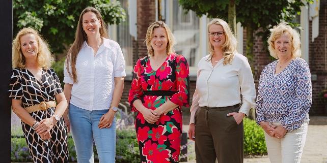 Vijf vrouwen poseren glimlachend voor het bedrijfspand van Notariskantoor Voorhout, omgeven door groene bomen en planten.